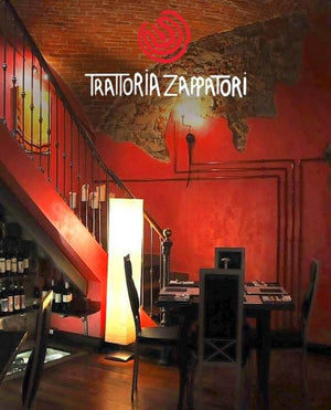 Interno del ristorante Trattoria Zappatori di Christian Milone. È visibile una scala a sinistra e svariate bottiglie di vino nel sottoscala. In primo piano si nota un tavolo con sedie nere e il tutto è illuminato da una luce calda.